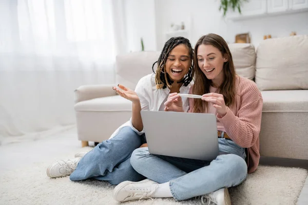Happy and lesbian woman showing pregnancy test near amazed african american girlfriend during video call on laptop — Stock Photo