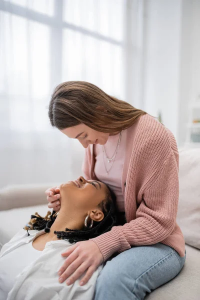 Heureuse et lesbienne femme assise sur canapé et regardant joyeuse petite amie afro-américaine — Photo de stock