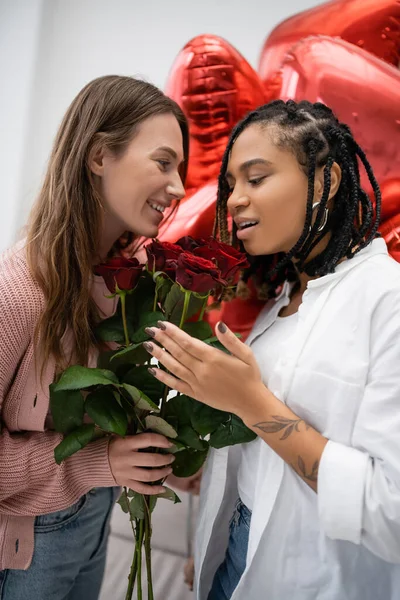 Sorrindo mulher lésbica segurando rosas vermelhas perto de namorada afro-americana espantada e balões no dia dos namorados — Fotografia de Stock