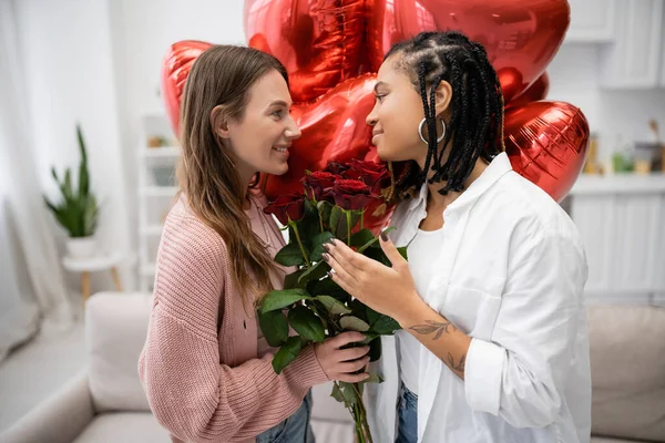 Улыбающаяся лесбиянка с красными розами и смотрящая на татуированную африканскую подружку рядом с воздушными шарами — стоковое фото