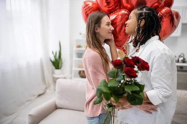Mulheres lésbicas inter-raciais olhando umas para as outras perto de rosas vermelhas e balões no dia dos namorados — Fotografia de Stock