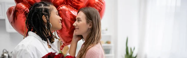 Donne lesbiche interrazziale che si guardano vicino a rose rosse e palloncini il giorno di San Valentino, striscione — Foto stock
