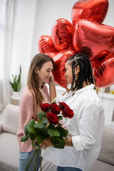 Lésbica afro-americana tocando bochecha de namorada com rosas vermelhas de pé perto de balões no dia dos namorados — Fotografia de Stock