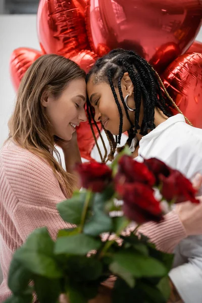 Mulheres lésbicas inter-raciais com olhos fechados perto de rosas vermelhas e balões no dia dos namorados — Fotografia de Stock