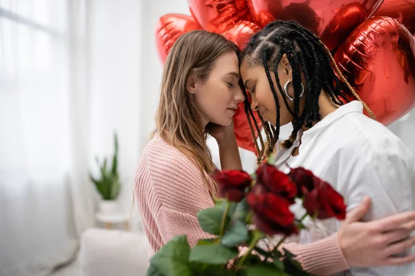 Mujeres lesbianas multiétnicas con los ojos cerrados cerca de rosas rojas y globos en el día de San Valentín - foto de stock