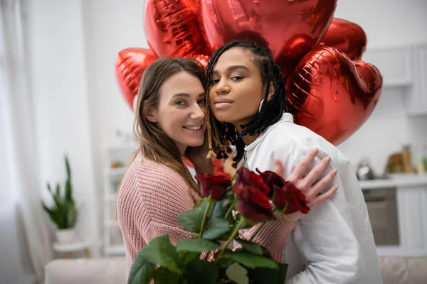 Mulheres lésbicas multiétnicas olhando para a câmera perto de rosas vermelhas e balões no dia dos namorados — Fotografia de Stock