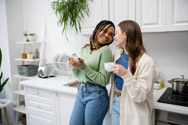Joyful and multiethnic lesbian women looking at each other while holding cups of coffee in kitchen — Stock Photo