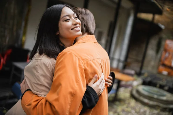 Smiling asian woman in trench coat hugging boyfriend on terrace of cafe — Stock Photo