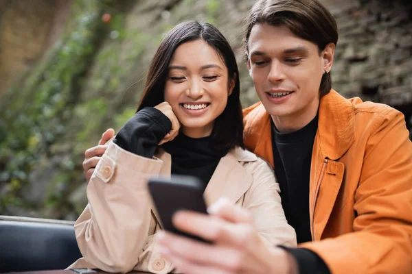 Young man hugging asian girlfriend in trench coat and using smartphone in outdoor cafe — Stock Photo