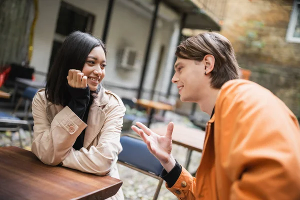 Sorridente donna asiatica in trench guardando fidanzato parlando in caffè all'aperto — Foto stock