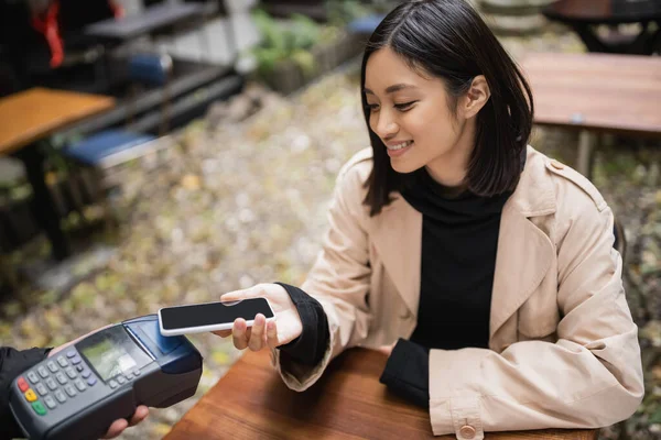 Sonriente mujer asiática en gabardina pagando con smartphone cerca de camarero sosteniendo terminal de pago en la terraza de la cafetería - foto de stock