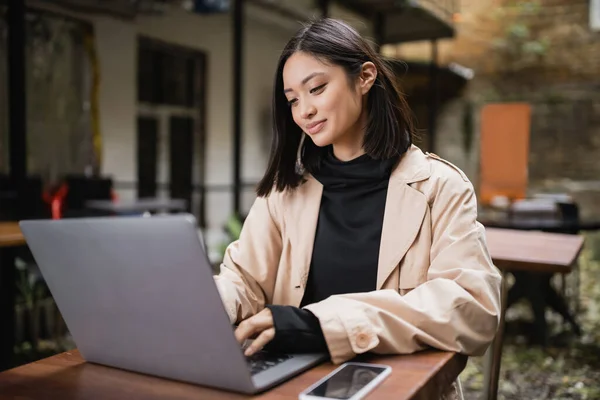Positivo asiático freelancer utilizando el ordenador portátil cerca de smartphone en la cafetería al aire libre - foto de stock