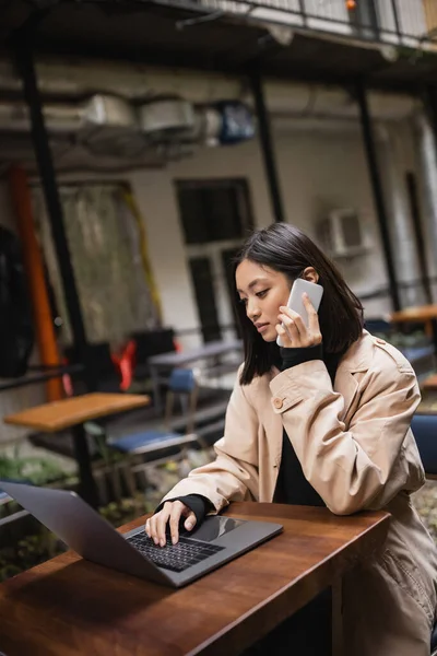 Jovem asiático freelancer em casaco de trincheira falando no smartphone e usando laptop no terraço do café — Fotografia de Stock