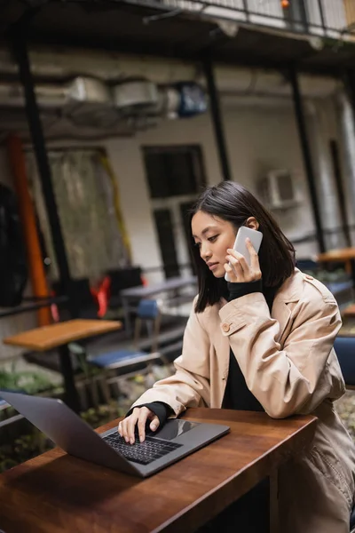 Young asian freelancer in trench coat talking on smartphone and using laptop on terrace of cafe — Stock Photo