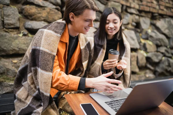 Lächelnder Mann in Decke mit Blick auf Laptop in der Nähe verschwommene asiatische Freundin mit Kaffee auf der Terrasse des Cafés — Stockfoto