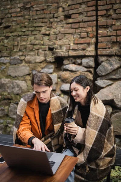 Cheerful asian woman holding coffee to go near boyfriend using laptop on terrace of cafe — Stock Photo