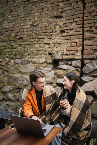 Multiethnisches Paar mit Decke und Kaffee unterhält sich in der Nähe von Geräten in Outdoor-Café — Stockfoto