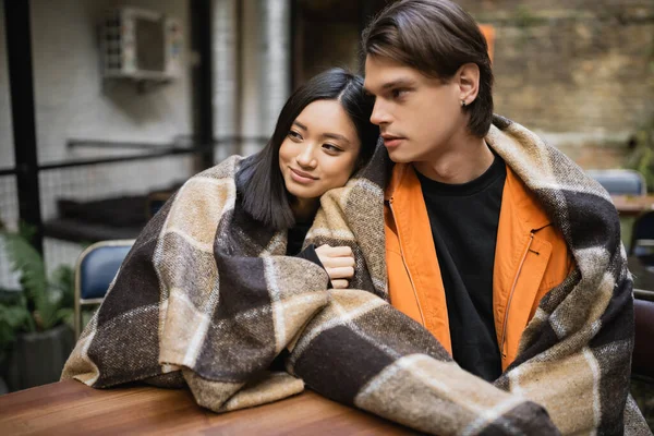 Smiling asian woman in blanket hugging boyfriend on terrace of cafe — Stock Photo