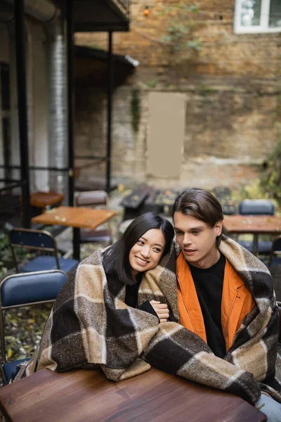 Cheerful asian woman in blanket hugging boyfriend on terrace of cafe — Stock Photo