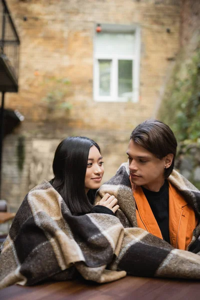 Young multiethnic couple in blanket looking at each other in outdoor cafe — Stock Photo