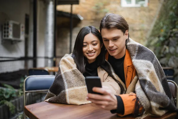 Smiling interracial couple in blanket using smartphone in outdoor cafe — Stock Photo