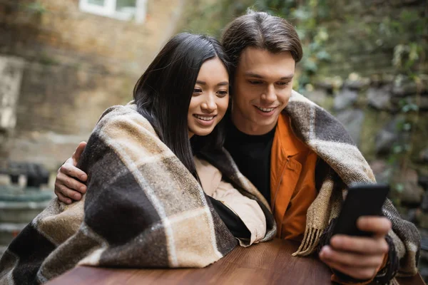 Young man hugging asian girlfriend in blanket while using blurred smartphone on cafe terrace — Stock Photo