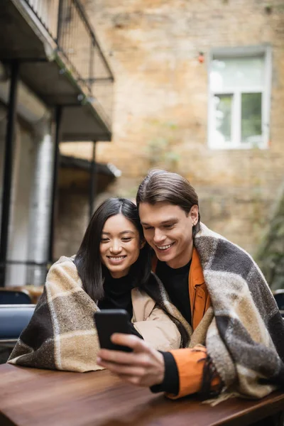 Positive interracial couple in blanket using cellphone on terrace of cafe — Stock Photo