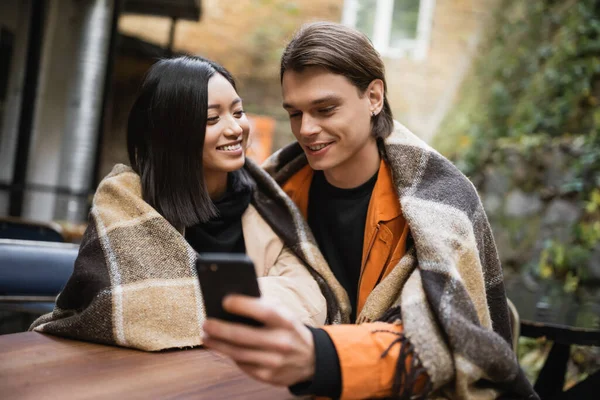 Улыбающаяся азиатка в одеяле смотрит на парня с размытым смартфоном в кафе на открытом воздухе — стоковое фото