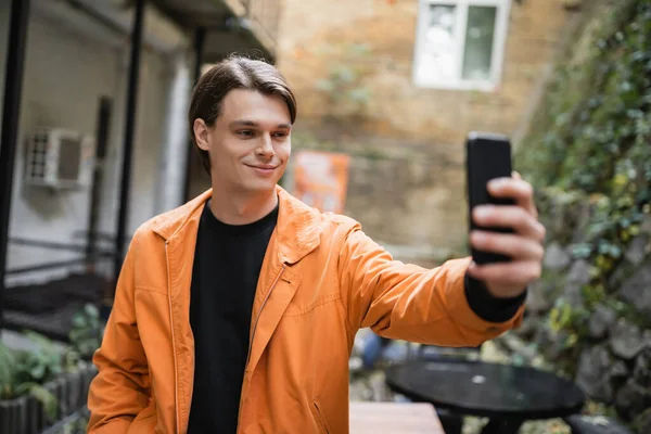 Smiling man in jacket taking selfie on smartphone on terrace of cafe — Stock Photo