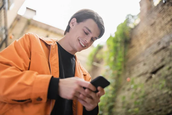 Low angle view of smiling man in jacket using mobile phone outdoors — Stock Photo