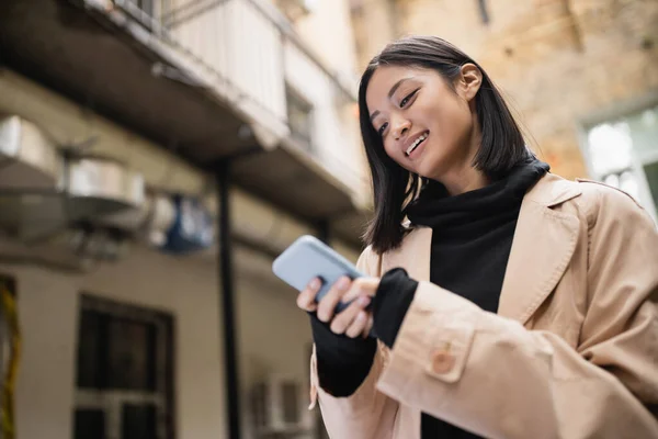Low angle view of smiling asian woman in trench coat using smartphone outdoors — Stock Photo