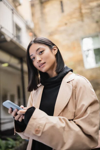 Stylish asian woman in trench coat holding smartphone and looking at camera outdoors — Stock Photo