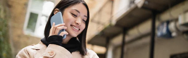 Positive asian woman in trench coat talking on smartphone outdoors, banner — Stock Photo
