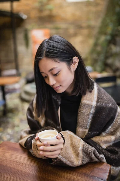 Jeune femme asiatique en couverture tenant tasse en papier avec cappuccino dans un café en plein air — Photo de stock