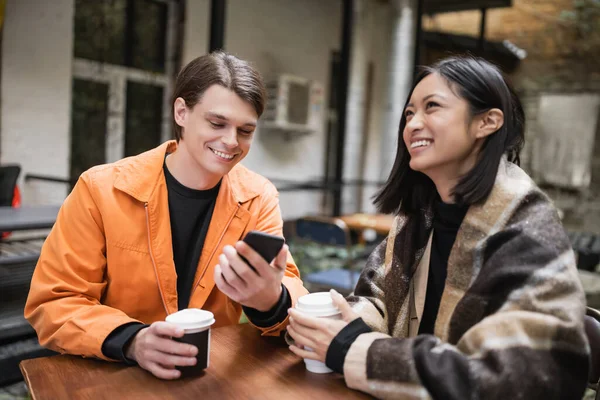 Positiver Mann benutzt Smartphone nahe asiatischer Freundin in Decke mit Kaffee auf Caféterrasse — Stockfoto