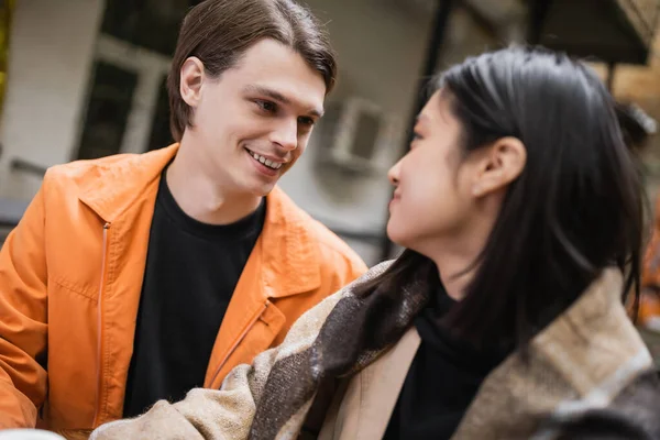 Smiling man looking at blurred asian girlfriend in blanket in outdoor cafe — Stock Photo