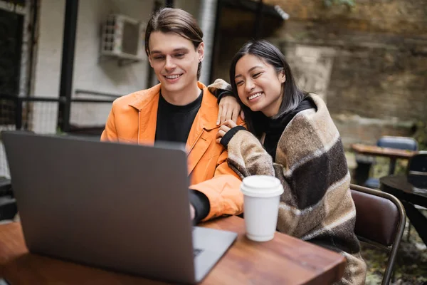 Smiling asian woman in blanket hugging boyfriend near laptop and coffee in outdoor cafe — Stock Photo