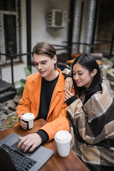 Positive asiatische Frau in Decke umarmt Freund mit Laptop in der Nähe von Kaffee auf der Terrasse des Cafés gehen — Stockfoto