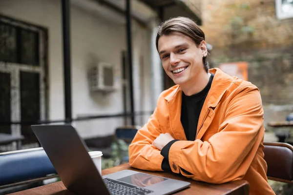 Young freelancer in jacket looking at camera near laptop and coffee to go on terrace of cafe — Stock Photo