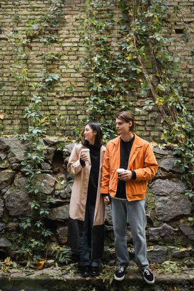 Молодая межрасовая пара держит кофе, чтобы пойти и держаться за руки возле стены на открытом воздухе — стоковое фото