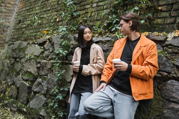 Young asian woman in trench coat holding paper cup near stylish boyfriend on urban street — Stock Photo