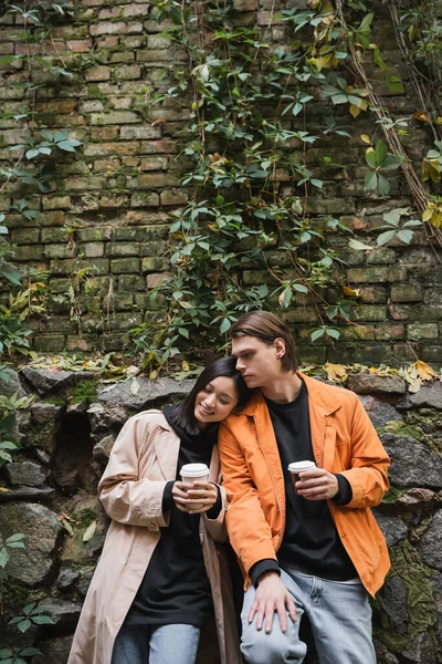 Souriant asiatique femme tenant à emporter boisson tout en se tenant près de petit ami sur la rue urbaine — Photo de stock