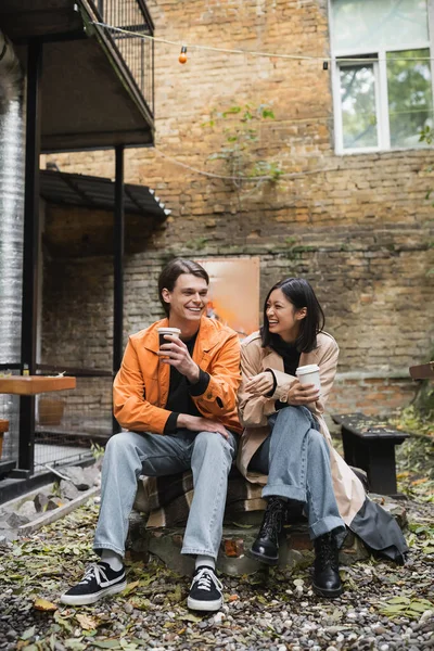 Cheerful and stylish multiethnic couple holding coffee to go on terrace of cafe — Stock Photo