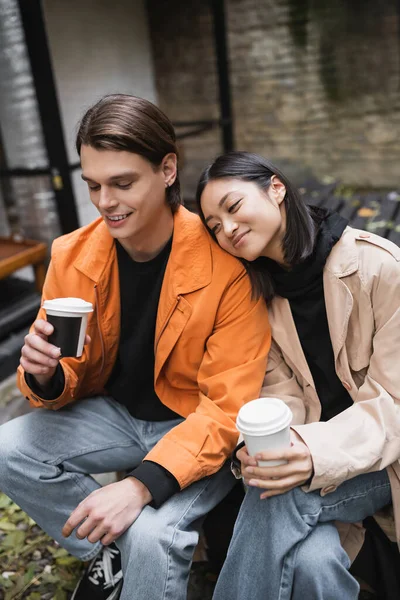 Усміхнений чоловік дивиться на каву, щоб підійти ближче задоволена азіатська дівчина в траншеї пальто у відкритому кафе — стокове фото
