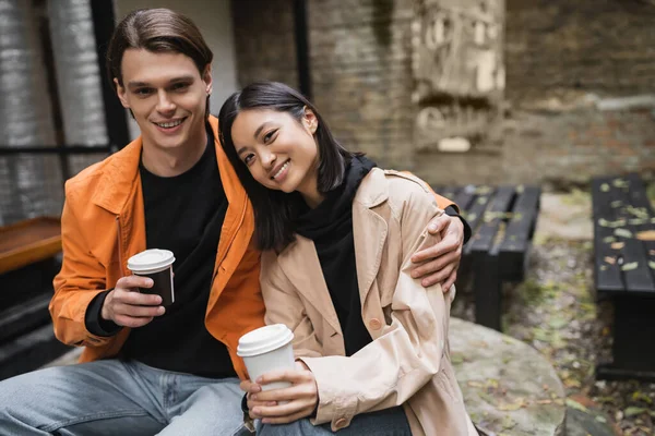 Positivo uomo tenendo tazza di carta e abbracciando ragazza asiatica in trench cappotto in caffè all'aperto — Foto stock