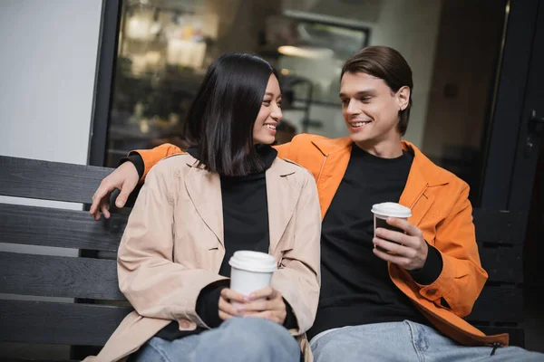 Усміхнена азіатка тримає каву, щоб підійти ближче до стильного хлопця на лавці у відкритому кафе — стокове фото