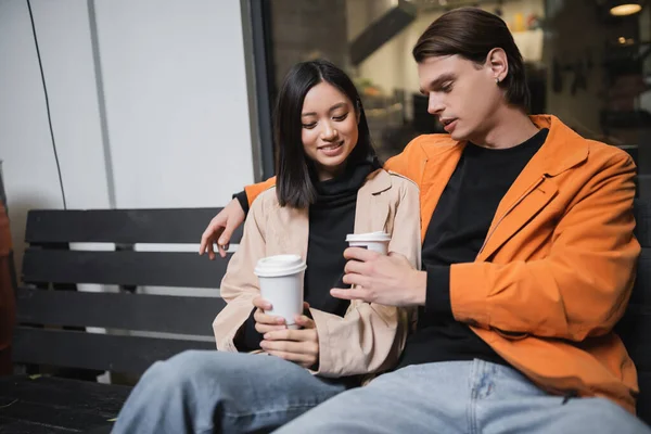 Молода міжрасова пара тримає каву, щоб піти, сидячи на лавці у відкритому кафе — стокове фото