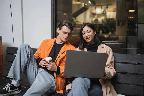 Smiling asian woman using laptop near boyfriend with paper cup on bench near cafe — Stock Photo