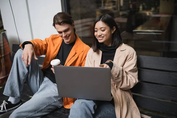 Sorrindo asiático mulher apontando para laptop perto namorado com café para ir no banco perto de café — Fotografia de Stock