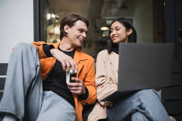 Smiling multiethnic couple with coffee to go and laptop looking at each other near cafe — Stock Photo